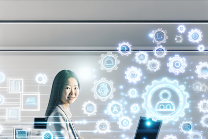 A woman in a gray blazer sits at a desk using a laptop with digital holographic icons surrounding her, depicting technology concepts.