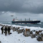 A snowy coastal scene with soldiers walking along a snow-covered path. A large cargo ship is sailing on the ocean, and several drones are flying overhead under a cloudy sky.
