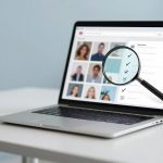 A laptop on a white table displaying a webpage with profile photos and checkboxes. A magnifying glass highlights the checkboxes.