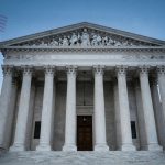The image shows the front facade of the U.S. Supreme Court building with its iconic columns and sculpted pediment. Two American flags are superimposed in the sky above. A large gavel is visible on a pedestal in the foreground. The sky is clear and blue.