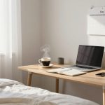 A tidy workspace with a wooden desk next to a bed. Items on the desk include a steaming cup of coffee, a closed laptop, a notebook, a smartphone, and a small potted plant. A calendar labeled "Day 1" is hanging on the wall. Sunlight filters through sheer curtains.