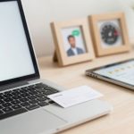 A laptop displaying a green open padlock icon on the screen sits on a desk. Nearby are a business card, a tablet showing a checklist with stars, framed photos, and a small potted plant.