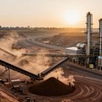A mining site at sunset with dusty conveyor belts, soil mounds, and industrial tanks. The sun sets in the hazy sky, creating a warm glow.