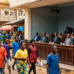 A bustling street market scene with people walking in colorful clothing, including blue, red, and yellow. On the right, a panel of officials in suits is seated at wooden desks, observing the scene. Market stalls with goods and vibrant umbrellas are visible in the background.
