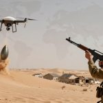 A soldier in camouflage and helmet aims a rifle at a flying drone dropping a bomb in a desert. Dust rises around the impact area, with scattered buildings in the background.