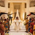 A bride in a white gown stands on steps in a large hall, flanked by guests in colorful African attire. Acrobats perform flips above, with a tall cake and sparklers in the background.