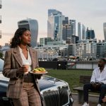 A woman in a plaid suit holds a coffee cup and food plate, standing by a luxury car. In the background, two people sit on a bench. The city skyline is visible at dusk.