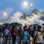 A group of people walking in the rain under umbrellas, with some taking photos. Streetlights illuminate the scene, and smoke fills the background.