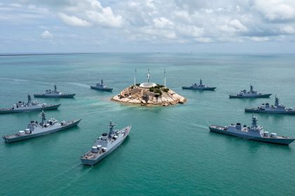 Seven naval ships encircle a small rocky island with a white monument and three flagpoles in the center of a calm turquoise sea under a cloudy sky.