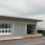 A laundromat with six front-loading washing machines and a vending machine outside. A white truck is parked nearby, and a green waste bin is on the sidewalk. The building has a metal roof and a garage-style door.