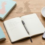 A wooden table with a teal book titled "Nelet Finance," an open notebook with handwritten notes and a pen, a smartphone displaying a website, and a cup of tea.