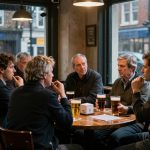 Seven people sit around a wooden table in a pub, discussing. They have pints of beer, and rain is visible outside the large windows.