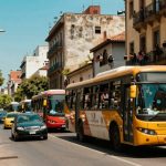 A busy street scene with a yellow bus and red bus in traffic. People are holding signs on the sidewalk, and others lean from balconies, holding pots. Buildings line the street under a clear blue sky.