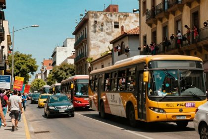 A busy street scene with a yellow bus and red bus in traffic. People are holding signs on the sidewalk, and others lean from balconies, holding pots. Buildings line the street under a clear blue sky.