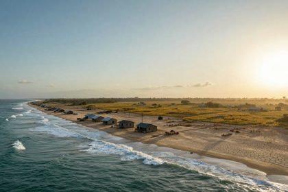 A coastal landscape at sunset, showing waves crashing on the shore. Small houses with blue roofs line the sandy beach. The sun is low in the sky, casting a golden light over fields and sparse vegetation in the background.