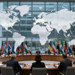 A group of people in suits sit around a large circular conference table with laptops and documents. A large world map is displayed on a glass wall behind them. Multiple national flags line the background, indicating an international meeting. The setting is a modern conference room with large windows.