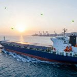 A cargo ship with solar panels on the deck navigates through the ocean at sunset. Cranes and a port are visible in the background.