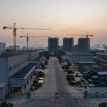 Aerial view of an industrial complex at dusk with construction cranes and high-rise buildings in the background. A wide, empty road runs between large factory buildings, with palm trees lining the sidewalks. The sky is a gradient of warm colors from the setting sun.