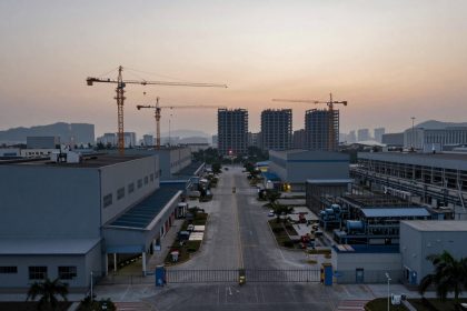 Aerial view of an industrial complex at dusk with construction cranes and high-rise buildings in the background. A wide, empty road runs between large factory buildings, with palm trees lining the sidewalks. The sky is a gradient of warm colors from the setting sun.