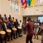 A group of people in colorful attire sing and play drums in a room with stained glass windows. Colorful banners hang from the ceiling.