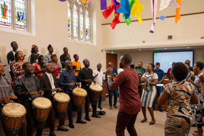 A group of people in colorful attire sing and play drums in a room with stained glass windows. Colorful banners hang from the ceiling.