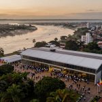 Aerial view of a modern building with a large gathering of people outside, surrounded by numerous flags. The building is near a river, with misty trees and city buildings in the background under a cloudy sky.