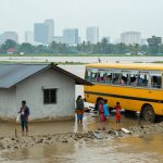 A flooded area with a yellow bus and a house partially submerged in water. People stand on the muddy ground; city buildings are visible in the background.