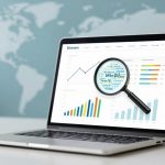 A laptop displaying graphs and charts with a magnifying glass overlay. Background features a blurred world map on a blue wall.