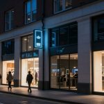A city street at dusk with people walking past a brightly lit clothing store. The store has large windows displaying various garments, and a glowing blue sign above. The building facade is modern with tall windows.
