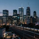 A cityscape at dusk featuring illuminated skyscrapers with various architectural designs. A train moves along the railway tracks in the foreground, and a few buildings have lit windows, adding to the urban ambiance. The sky is a deepening blue as evening approaches.