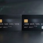 Two black debit cards with gold chips displayed side by side on a dark surface. Cards have embossed numbers and text.