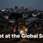 A group of people sit around a lantern at dusk in a rural area, with a city skyline in the background. The text "Debt at the Global South" overlays the image.
