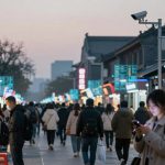 Busy street market at dusk with people walking and shopping. Vendors serve food under bright signs, surveillance cameras overhead.
