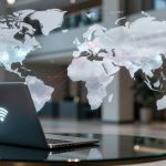 Laptop on a glass table displays a glowing Wi-Fi symbol, with a transparent world map overlay showing global network connections. Office setting in the background.