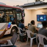 A man holds a smartphone while using a laptop, sitting outside a bus. Nearby, three people work on laptops under a shelter with screens displaying data.