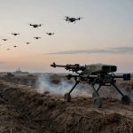A robotic device with wheels and a mounted gun is positioned in a trench on a battlefield. Multiple drones fly overhead, and tanks are visible in the background under a cloudy sky at sunset.