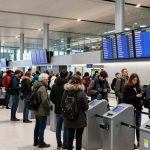People stand in line at an airport check-in area. They wear winter clothing and carry luggage. Flight information screens are visible above.