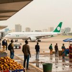 A white airplane with green accents taxis on an airport tarmac. In the foreground, vendors sell produce under colorful tents. People are walking and observing the scene. A large flag is visible on the left, and city buildings are in the background.