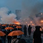 Crowd holding orange umbrellas walking down a city street with fires burning in the background, producing thick smoke. Tall buildings are visible under a dusky sky.