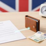 A desk with a UK flag backdrop holds a document, a brown wallet, foreign currency, a small clock, and a mini shopping basket with apples.