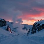 Snow-covered mountain pass at sunset, with silhouettes of people carrying skis on a ridge. Sky is pink and orange, with clouds.