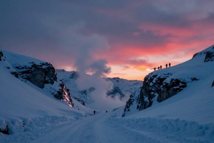 Snow-covered mountain pass at sunset, with silhouettes of people carrying skis on a ridge. Sky is pink and orange, with clouds.