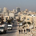 A cityscape with mid-rise buildings in the background. In the foreground, trucks drive down a street lined with beige stone buildings. Rubble is visible on the right, with people walking among the debris.