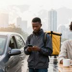 A man with a yellow delivery backpack looks at his phone beside a parked car. Another person cleans the car window with a cloth. A woman sits at an outdoor table using a laptop, with a mug nearby. Cityscape with skyscrapers in the background.