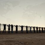 A group of people stand side by side, with hands raised, holding onto a tall fence. The sky is a pale gradient, and a faint world map overlays the background. Birds are visible in the sky.