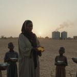 A woman in a headscarf holds a handful of grain, surrounded by four children with metal bowls. They stand on cracked, dry ground with silos and a setting sun in the background.