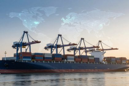 A large cargo ship loaded with multicolored shipping containers is docked at a port with cranes. A world map overlay is visible in the sky.