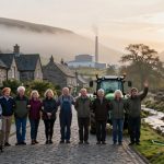 A group of eleven people stands on a cobblestone street in front of stone cottages. They are smiling, with one person waving. A tractor is parked behind them. The setting is rural with a stream, grassy areas, and hills in the background. The sun is setting, creating a warm glow.