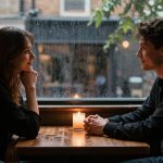 A man and woman sit across from each other at a wooden table in a dimly lit cafe. A lit candle is in the center. Rain falls outside the window behind them. They are smiling and making eye contact.