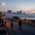 People walk along a waterfront street at dusk, with silhouetted skyscrapers in the background. Streetlights illuminate the scene, and graffiti covers a low wall by the water. Smoke or fog partially obscures the distant buildings.